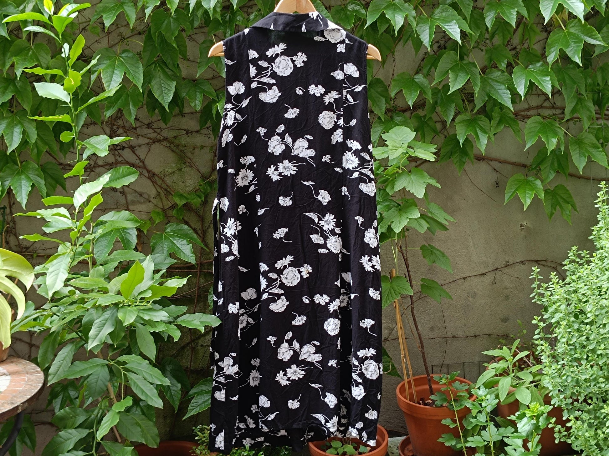 A close-up shot of the back of a black vintage sleeveless dress with white floral patterns. The vintage dress is hanging from a clothing line outdoors in a garden and the background is green and leafy