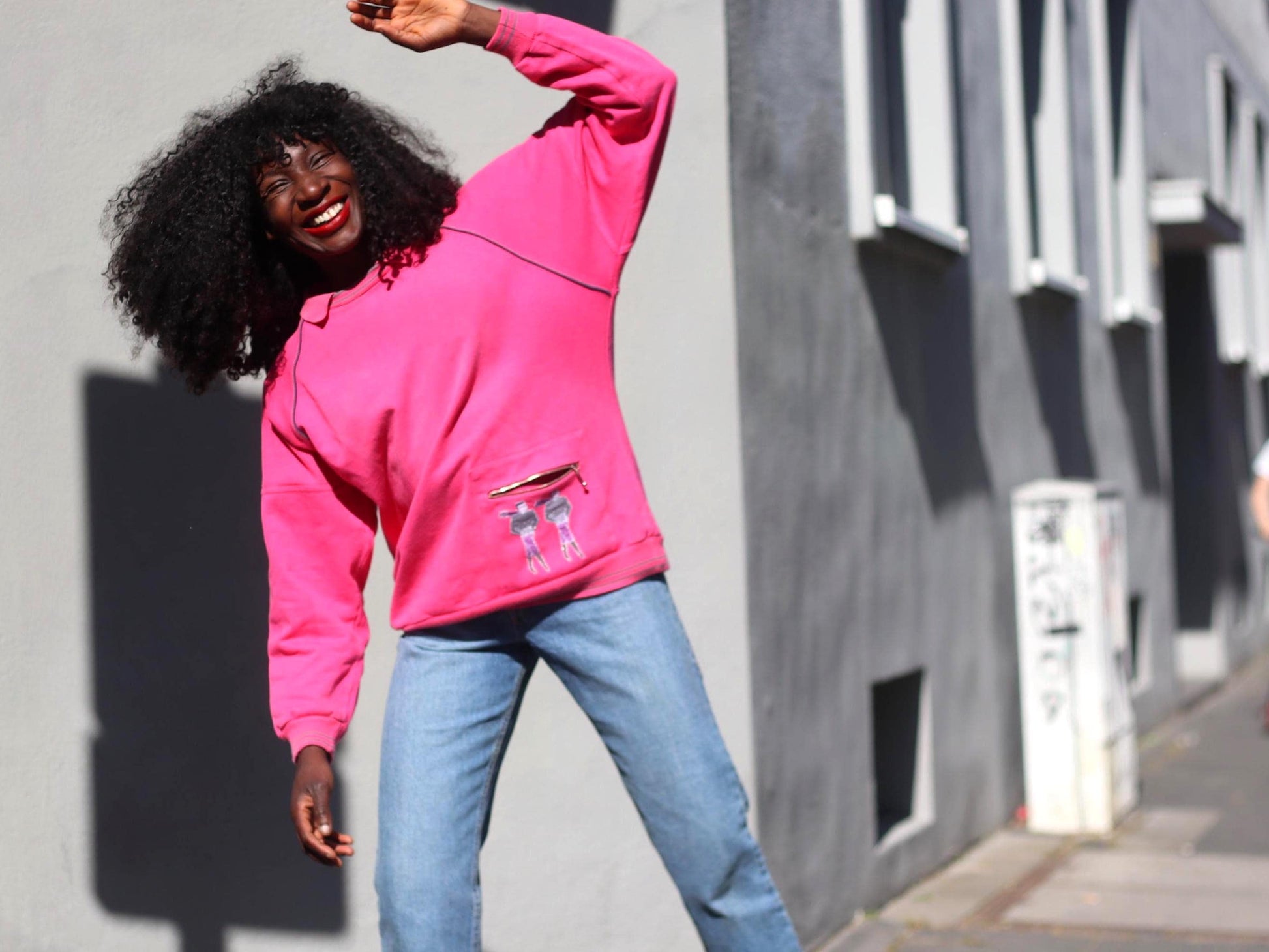 Female Model with curly black hair wearing a bright pink 80s vintage sweatshirt with a front zip pocket and cartoon graphic in front of a grey wall on a sunny day.