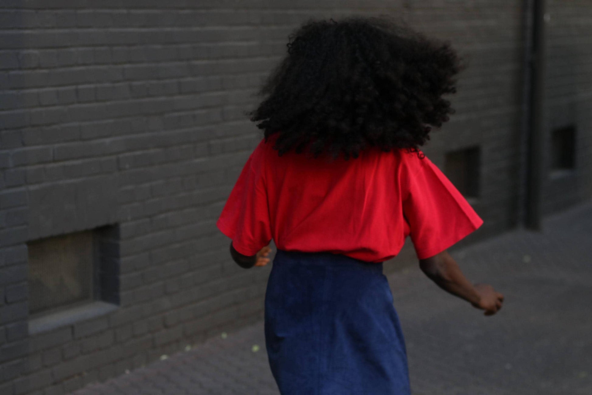 Back view  portrait of a female  person with dark, curly hair, wearing a red collared shirt and a dark blue skirt with a zipper detail, photographed against a textured gray wall.