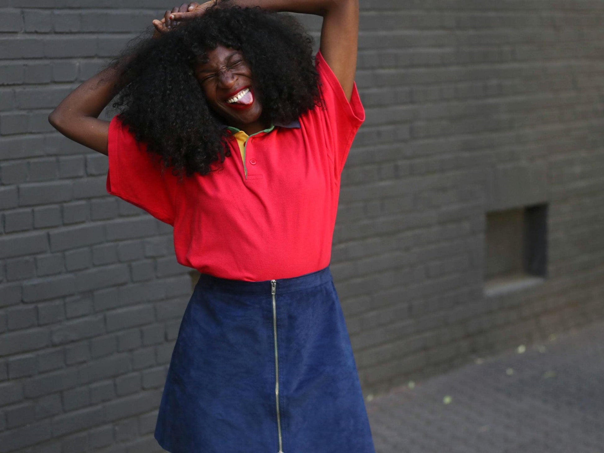 A smiling model with dark, voluminous curly hair is playfully posed with their hands near their head, wearing a bright red collared polo and a vintage dark blue suede skirt with a front zipper, against a textured gray brick wall.