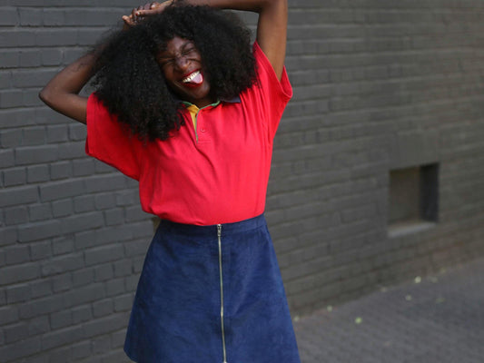 A smiling model with dark, voluminous curly hair is playfully posed with their hands near their head, wearing a bright red collared polo and a vintage dark blue suede skirt with a front zipper, against a textured gray brick wall.
