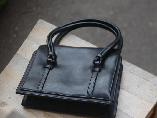 A black vintage 1970s leather compact handbag with double top handles and gold-tone hardware, resting on a light wooden surface.