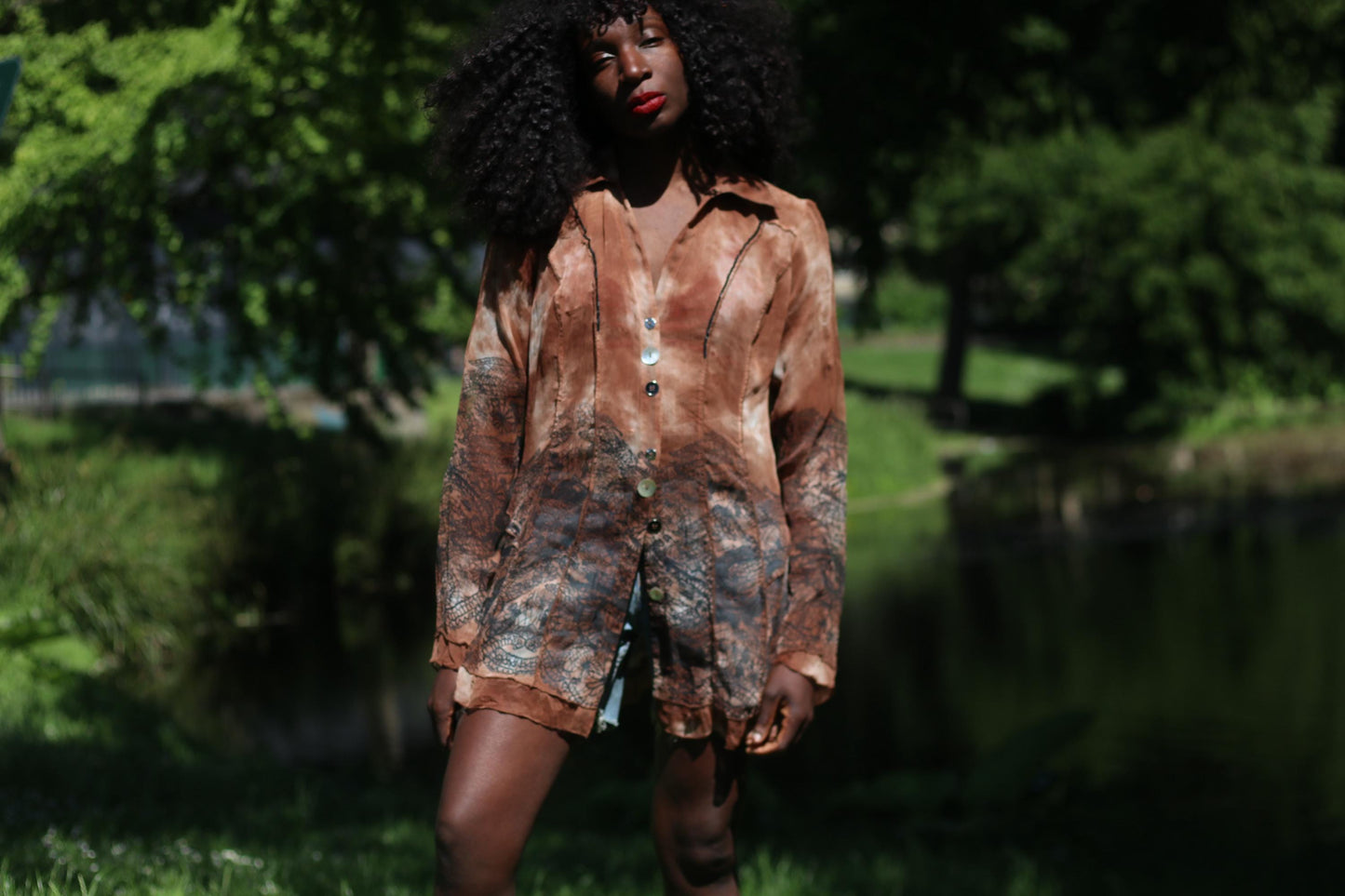 Female model with curly black hair in a BibA vintage brown blouse with leaf motif and lightweight mesh fabric styled with blue denim shorts standing outdoors with a lake in the background