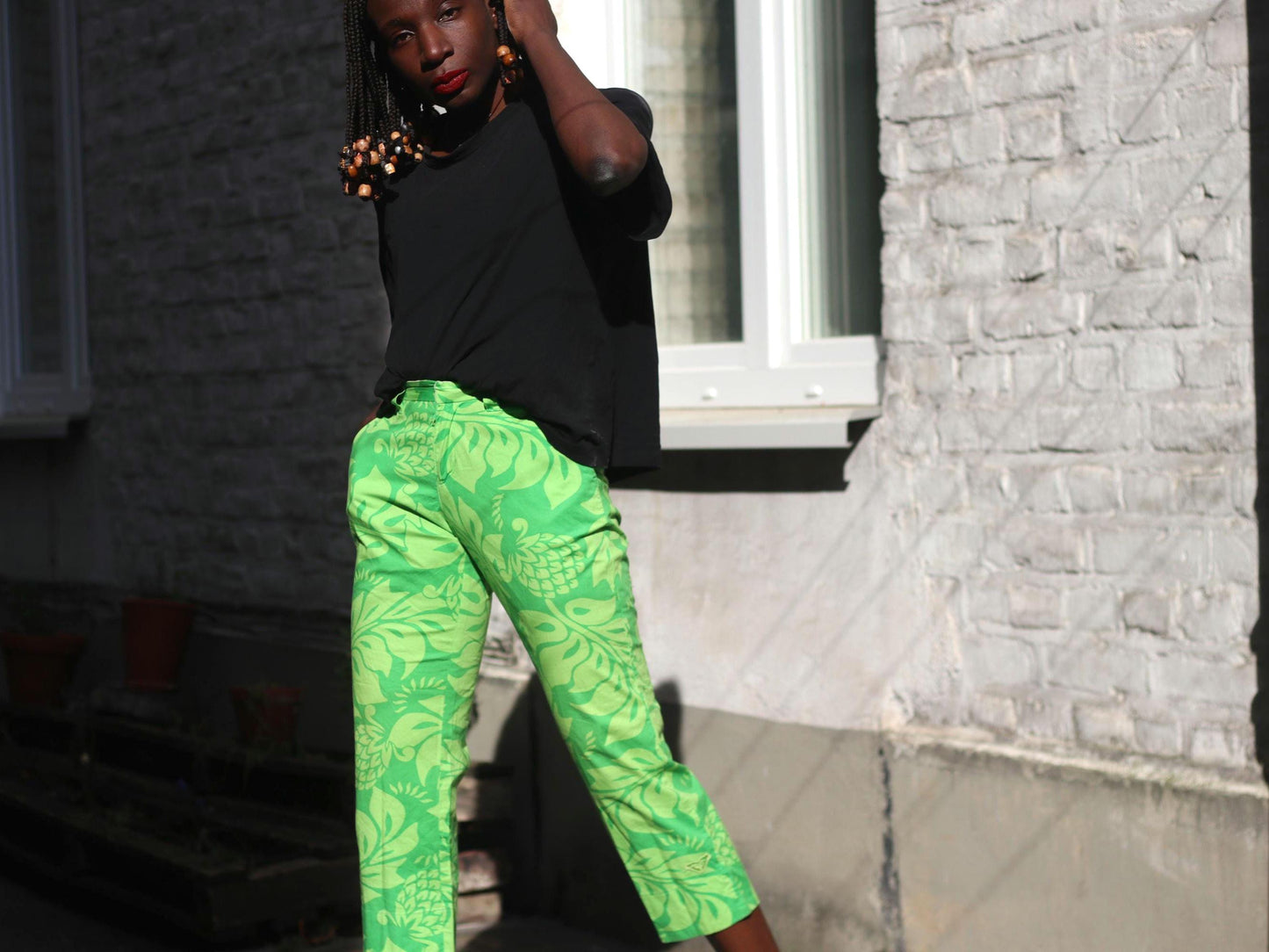 Dark-haired female with beaded braids wearing in vibrant green floral capri pants by Quiksilver stands outdoors by a building.