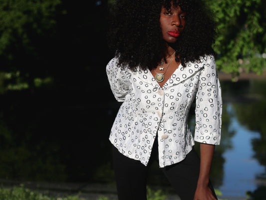 A female model with curly black hair wearing a white vintage blouse with a black abstract circle print and black pants, standing outdoors by water and trees.