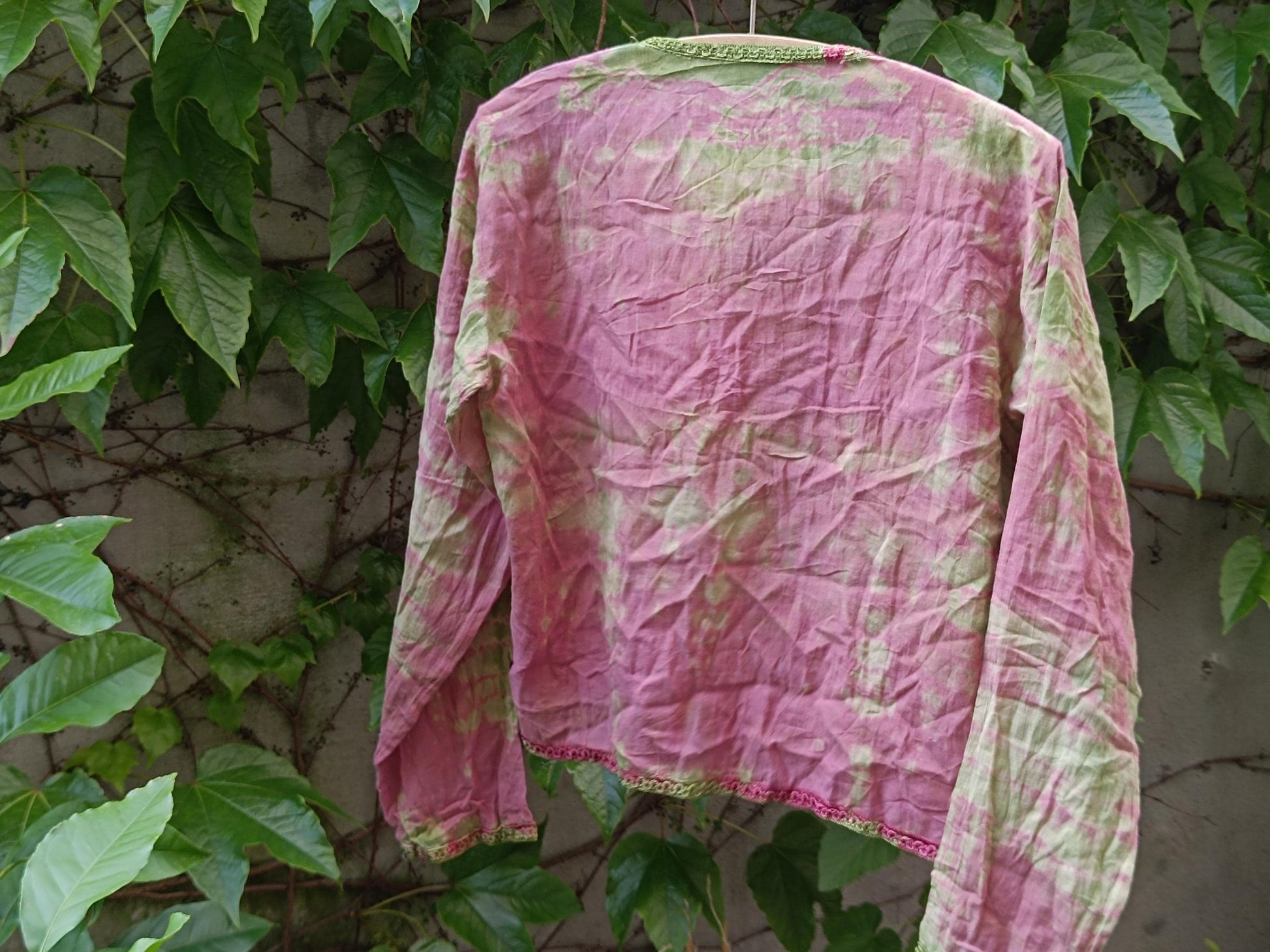 Back view of a vintage boho chic tie-dye  blouse with pink and green colors photographed hanging outdoors on a clothing line with leaves in the background. The still life image showcases the print and texture of the retro long-sleeved blouse.
