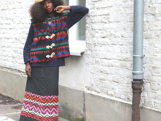 Femal model wearing a  vintage black knit maxi skirt with a wide, colorful geometric pattern band at the bottom hem photographed outdoors against a white brick exposed wall