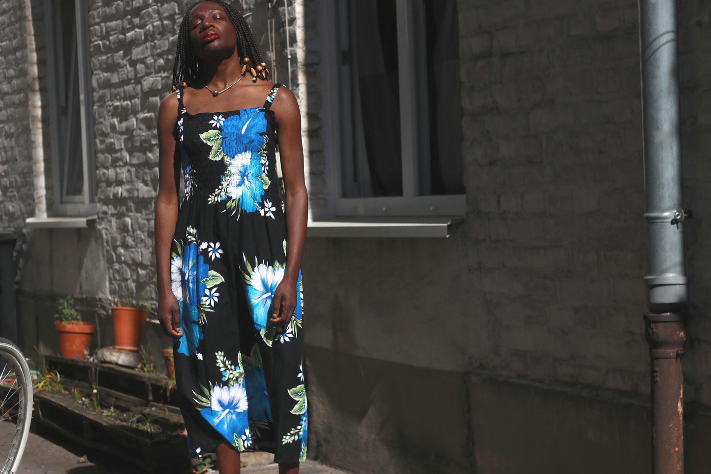 A female model of African heritage with dark hair styled in braids with brown beads at the ends, models a vintage sleeveless, flowing rayon summer dress with a black base and large, vibrant blue and white floral prints, standing outdoors against a textured stone building with windows and potted plants.