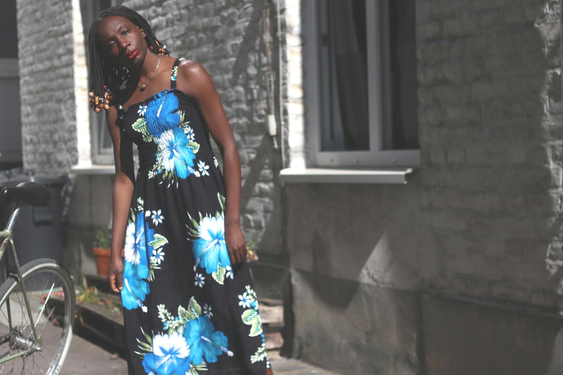 A sleeveless rayon summer dress with a predominantly black background and striking, large blue and white floral designs is showcased on a Black model standing in an artistic fashion. The dress has a relaxed fit and falls to the ankles, and the setting includes a stone building with visible windows and some