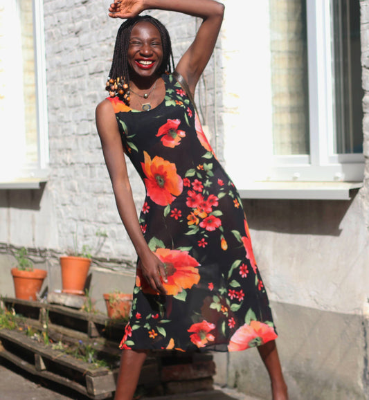 A female model of African heritage wearing a black semi-sheer vintage sleeveless midi dress with a red and orange floral print is photographed outdoors. She smiles broadly, with their arms raised above their head. The dress features large red and orange flowers against a black background. The person is accessorized with a  necklace. The background shows a white windowsill, a brick wall, terracotta pots, and a wooden pallet with plants.