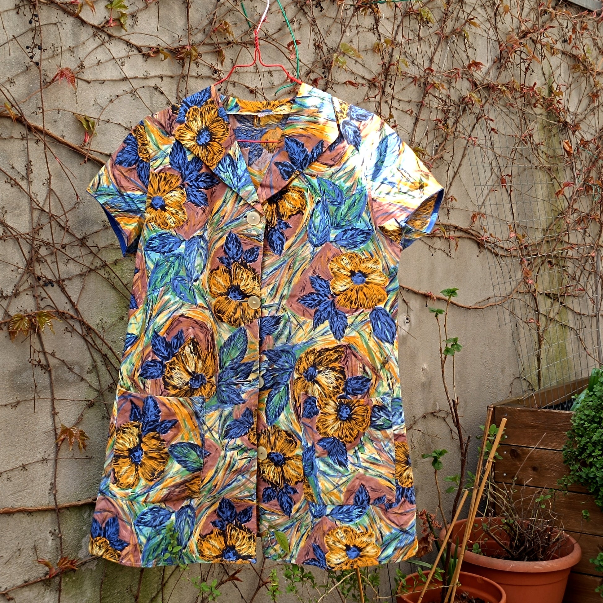 70s Vintage floral print tunic dress with a blue and yellow palette, captured outdoors  hanging on a hanger in a garden against a wall with browning twines, glowing in soft light with a nostalgic 70s feel.