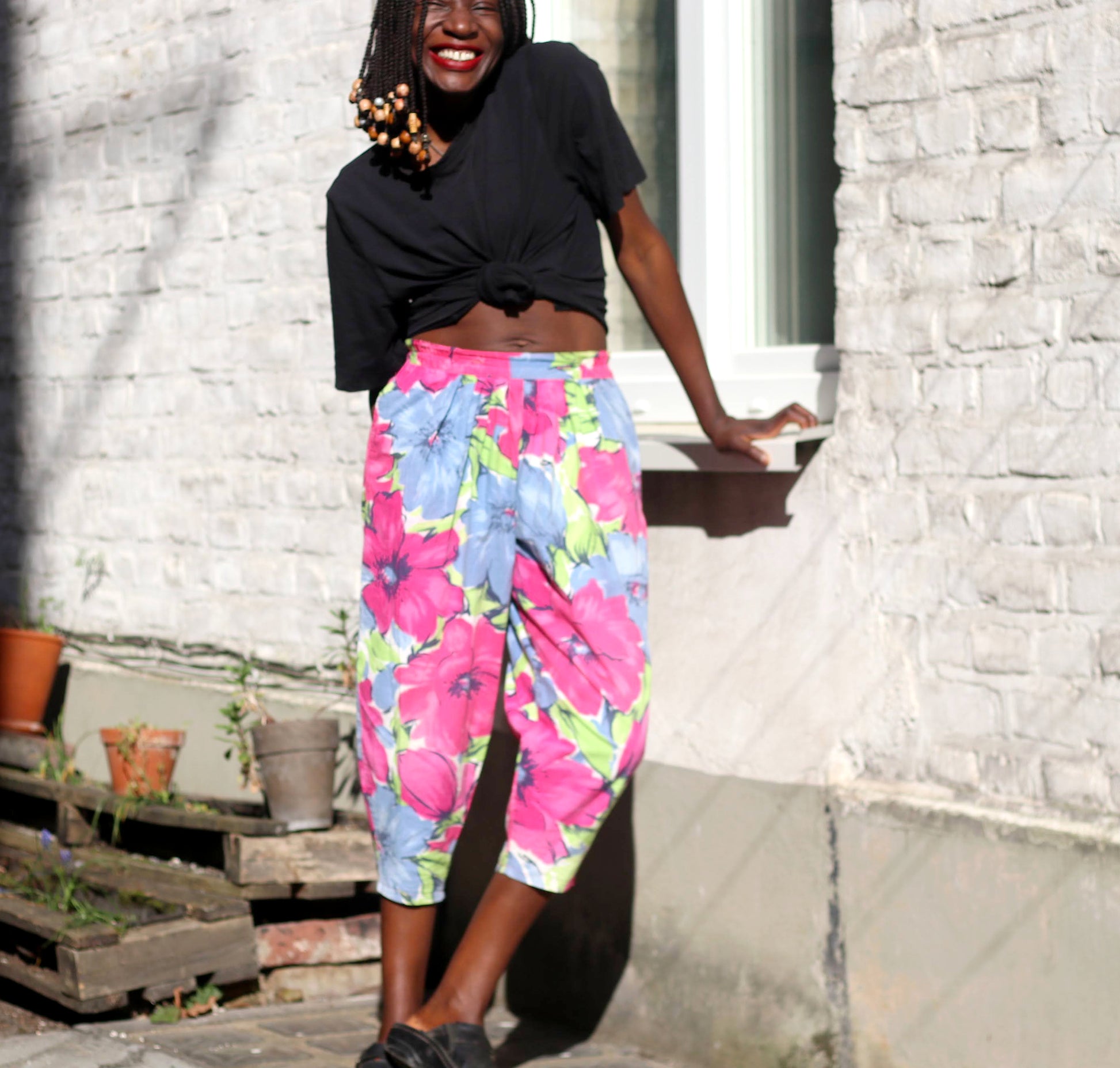 A smiling model with braids in a pair of vintage 90s  tropical floral-print summer capri pants poses against a sunlit brick wall.