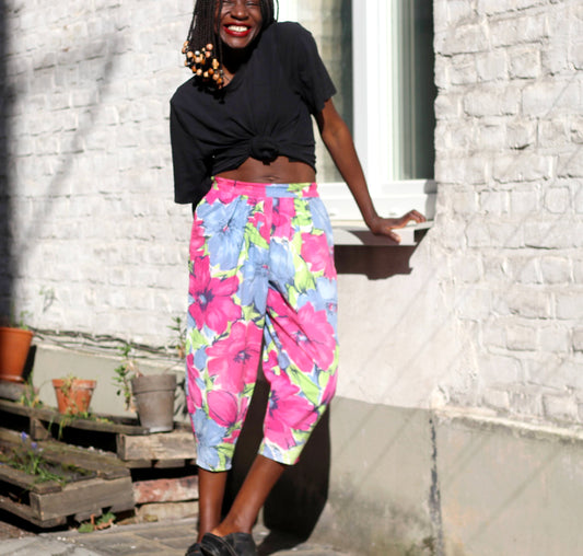 A smiling model with braids in a pair of vintage 90s  tropical floral-print summer capri pants poses against a sunlit brick wall.