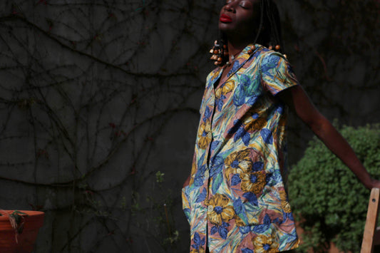 Woman in a vintage floral print tunic dress with a blue and yellow palette, captured outdoors in a garden against a wall with browning twines, glowing in soft light with a nostalgic 70s feel.