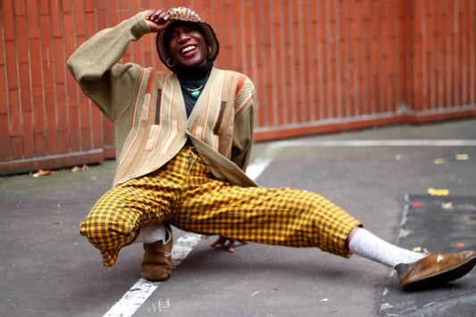A fashionable black woman crouched down infront of a brick wall one knee bent and one leg straight. One hand is on her animal print hat. She is wearing a black turtleneck. a brown striped sweater. Her trousers are yellow with a checkered print