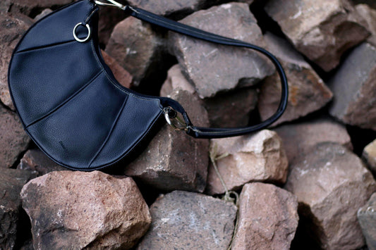 A 90s minimalist designer handbag in black with chic silver detail is photographed against a backdrop of brown stones.Thedesigneris Daniel Ray
